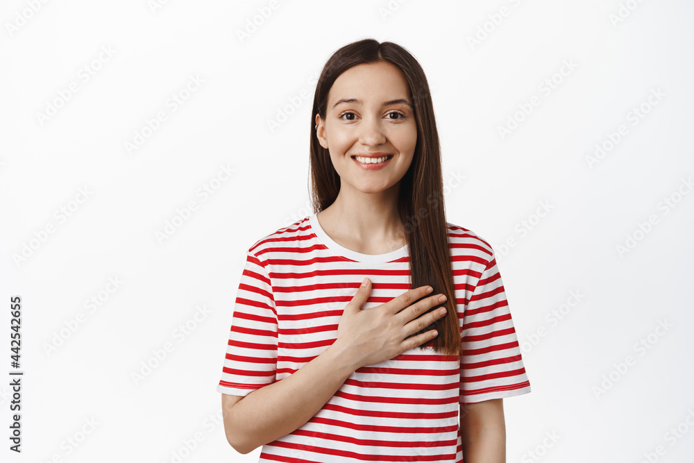 Image of young brunette woman hold hand on heart and smiling, with deep thoughts in her mind, look thankful or delighted, flattered with smth, standing against white background Image of young brunette woman hold hand on heart and smiling, with deep thoughts in her mind, look thankful or delighted, flattered with smth, standing against white background