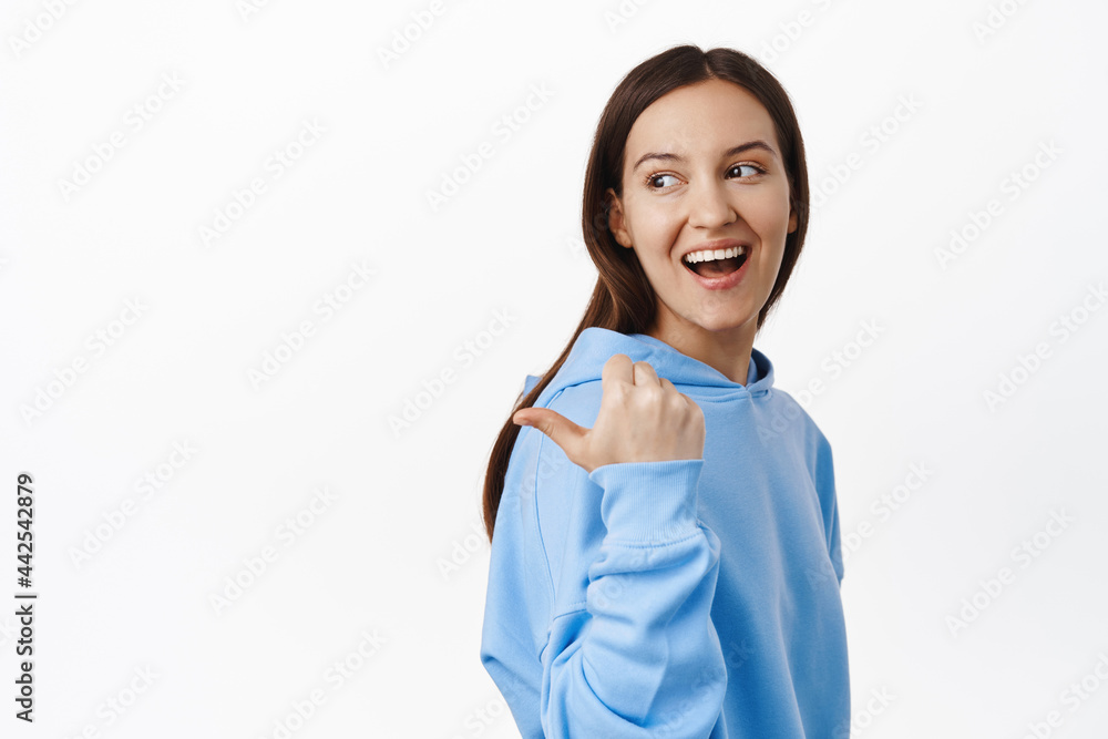 Portrait of young candid girl, smiling happy, pointing left and looking behind her shoulder at sale banner, standing in hoodie against white background Portrait of young candid girl, smiling happy, pointing left and looking behind her shoulder at sale banner, standing in hoodie against white background