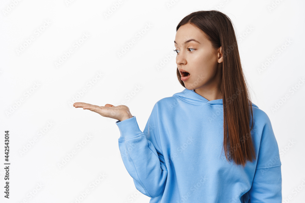 Portrait of brunette girl 20s years, looking at her palm, empty open hand, holding smth, display at item, advertisement of product on sale, standing against white background Portrait of brunette girl 20s years, looking at her palm, empty open hand, holding smth, display at item, advertisement of product on sale, standing against white background
