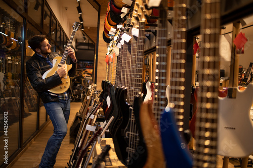 Proud young rocker musician in leather jacked playing electric guitar in music shop.