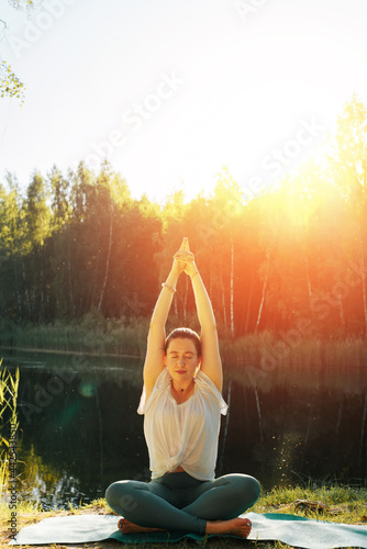 Attractive, athletic young woman practicing yoga asana outdoors. Girl yogi sitting onsports mat pose Sama Asana Sukhasana, hands top of Sat Kriya position. Yoga and meditation at sunset or sunrise.