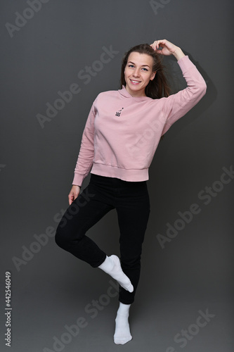 Full length studio portrait of cute caucasian girl in pink sweater and black pants with smile posing on gray background