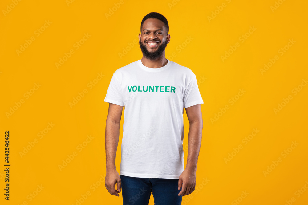 Happy Black Volunteer Guy Standing Wearing Uniform T-Shirt, Yellow Background Happy Black Volunteer Guy Standing Wearing Uniform T-Shirt, Yellow Background
