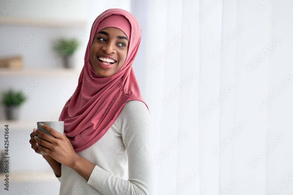 Happy Black Muslim Woman In Hijab Drinking Coffee Near Window At Home Happy Black Muslim Woman In Hijab Drinking Coffee Near Window At Home