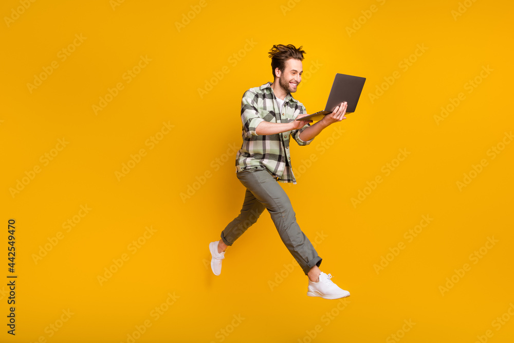 Full body profile side photo of young man happy positive smile jump use laptop isolated over yellow color background Full body profile side photo of young man happy positive smile jump use laptop isolated over yellow color background