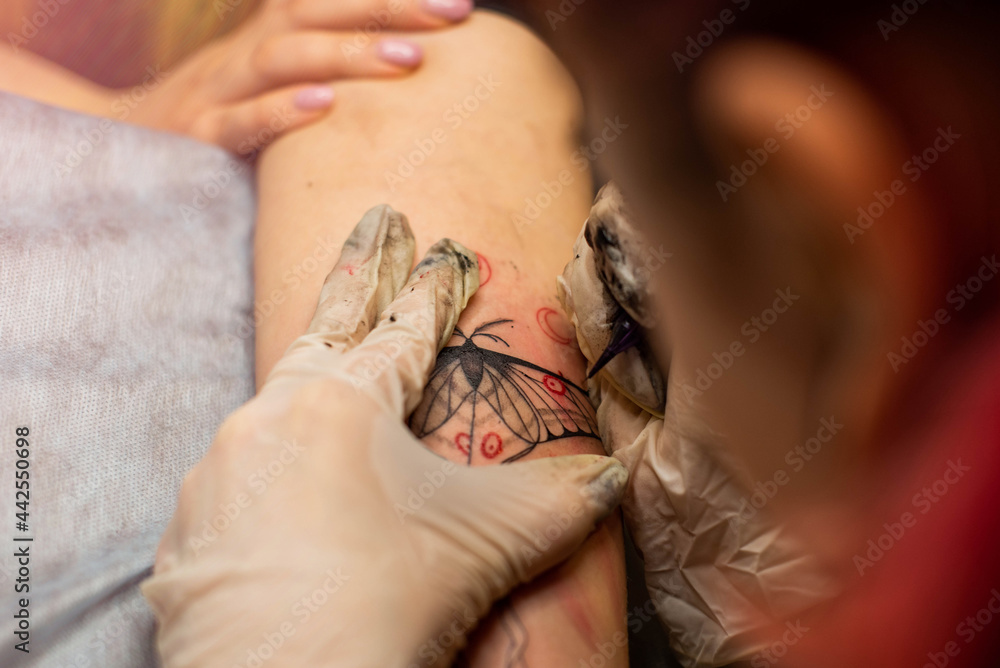 Obraz premium Master tattoo draws the red paint on the clients tattoo. Tattoo artist holding a pink tattoo machine in black sterile gloves and working on the professional blue mat.