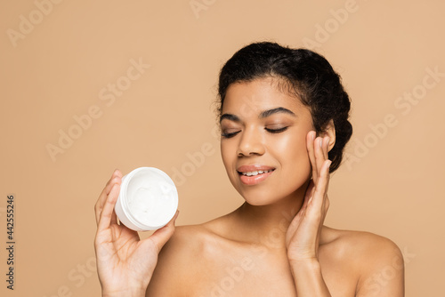 smiling african american woman with bare shoulders looking at container with cosmetic cream isolated on beige