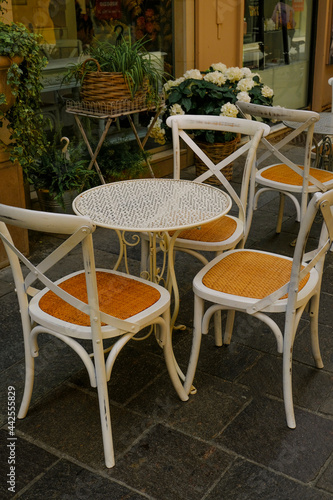 White wooden table with chairs across plants in pots in restaurant terrace. Cafe exterior. City life