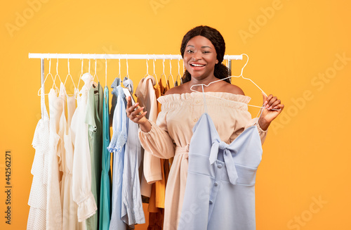 Happy black woman using cellphone for internet shopping, picking dress near clothing rail, buying summertime wardrobe