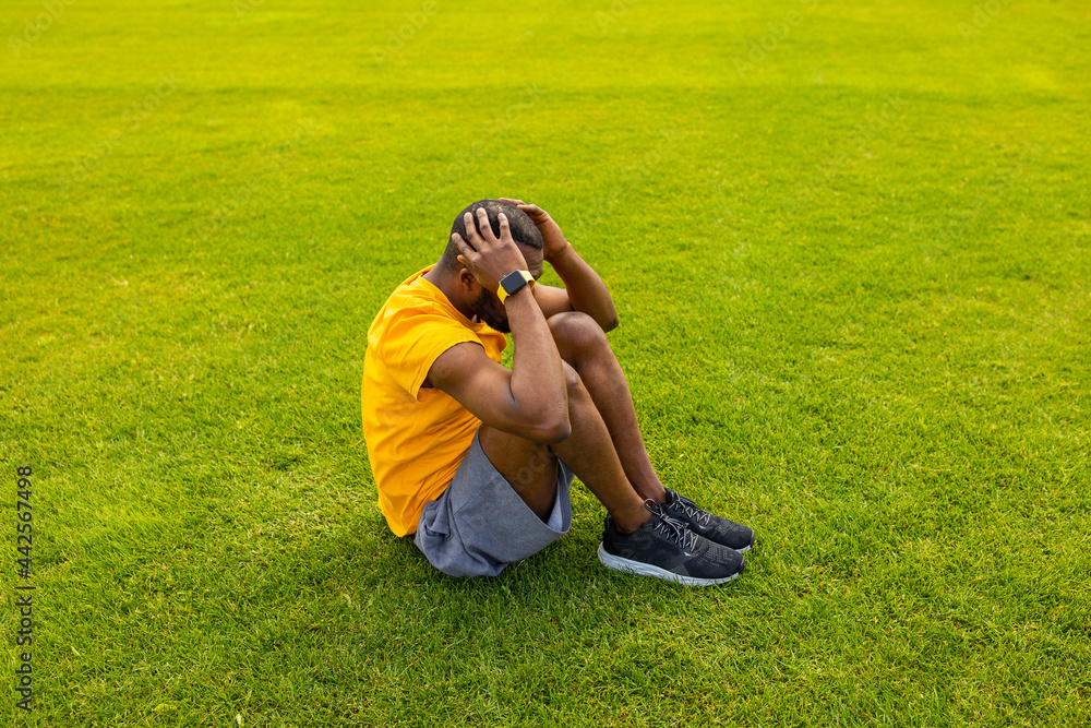Active Afro American male in trendy yellow sportswear, sitting on the grass doing crunch exercises at stadium outdoors, millennial black male holding head and doing abs workout on fresh air. Active Afro American male in trendy yellow sportswear, sitting on the grass doing crunch exercises at stadium outdoors, millennial black male holding head and doing abs workout on fresh air.