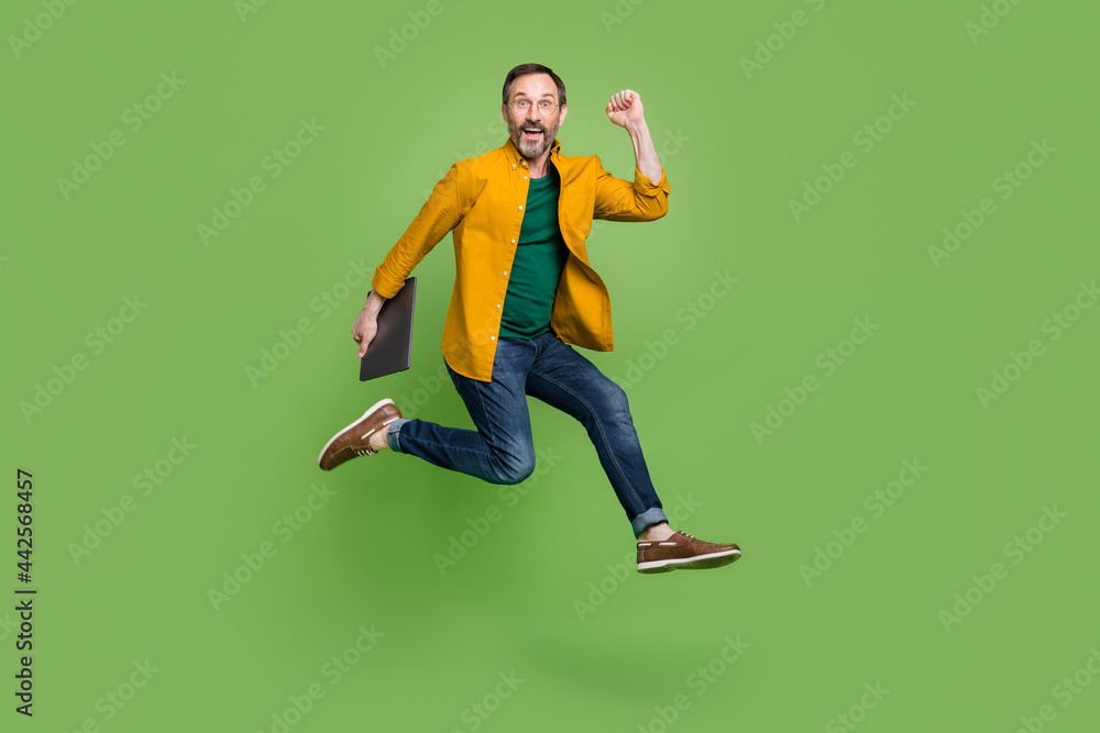 Full size profile side photo of mature business man happy positive smile jump up hold laptop isolated over green color background Full size profile side photo of mature business man happy positive smile jump up hold laptop isolated over green color background