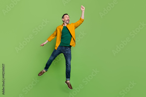 Full size photo of handsome mature man happy positive smile try to catch umbrella jump isolated over green color background