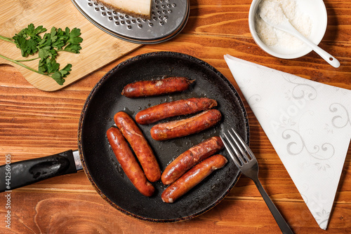 Tasty fried sausages in frying pan with Parmigiano and parsley, over wooden background. Top view