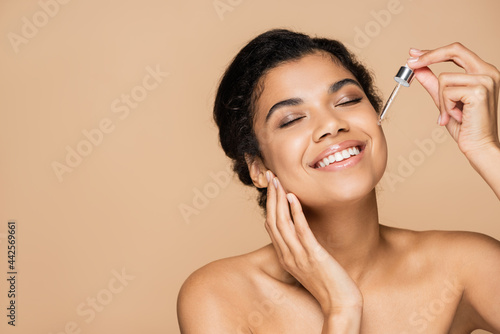 happy african american woman with bare shoulders applying serum isolated on beige
