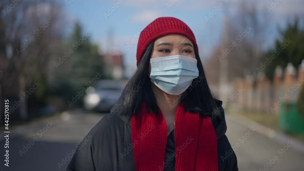 Front view portrait of young Asian woman in Covid face mask strolling outdoors on sunny spring autumn day. Relaxed millennial walking in slow motion enjoying leisure on coronavirus quarantine Front view portrait of young Asian woman in Covid face mask strolling outdoors on sunny spring autumn day. Relaxed millennial walking in slow motion enjoying leisure on coronavirus quarantine