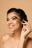joyful african american woman with bare shoulders applying mascara isolated on beige