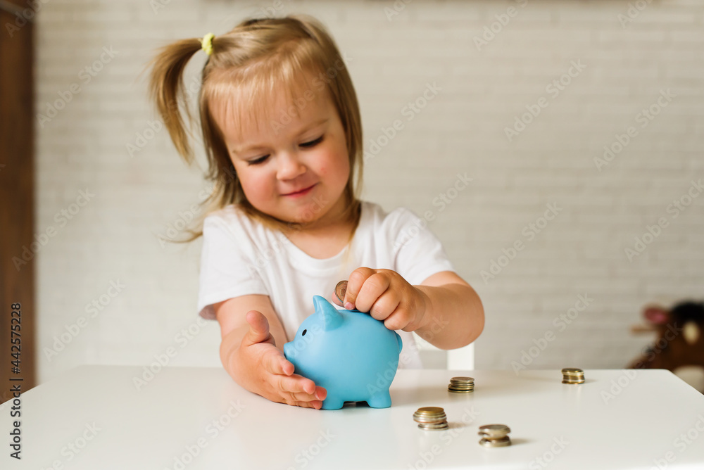Little Caucasian girl putting coins in to the pig-shaped money box, blue piggy bank. The child manages and deposits his finance, girl saving money in a piggybank for future. Little Caucasian girl putting coins in to the pig-shaped money box, blue piggy bank. The child manages and deposits his finance, girl saving money in a piggybank for future.