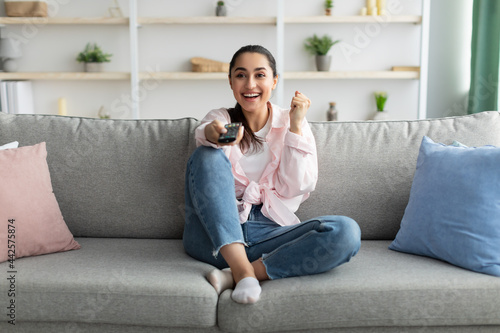 Excited woman watching tv shaking clenched fist holding remote control