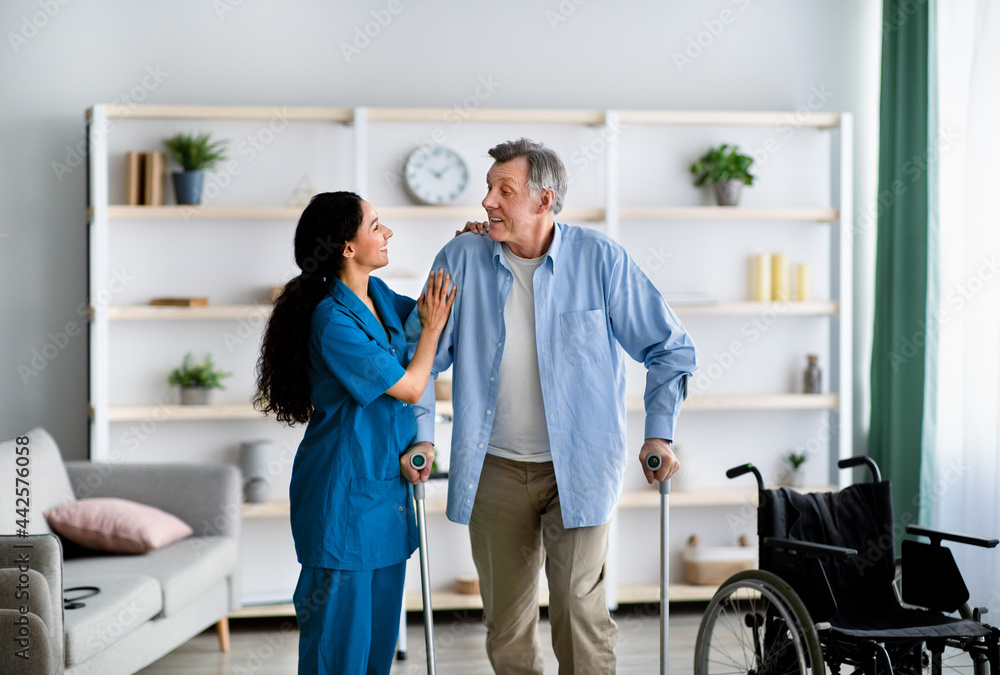 Obraz premium Female nurse helping elderly male to walk with crutches at home. Professional medical care for disabled older patients