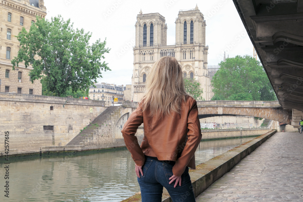 Blonde woman in trousers, standing from behind on the banks of the banks of the Seine. Cathedral of Notre Dame de Paris deliberately blurred in the background.