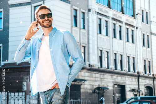 Handsome smiling stylish hipster lambersexual model.Modern man dressed in blue shirt. Fashion male posing on the street background in sunglasses. Outdoors at sunset. Using smartphone.Talking © halayalex