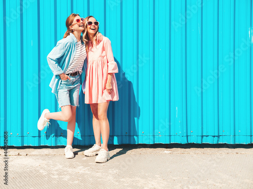 Two young beautiful smiling hipster female in trendy summer clothes and dress.Sexy carefree women posing in the street near blue wall.Positive pure models having fun at sunset, hugging and going crazy © halayalex