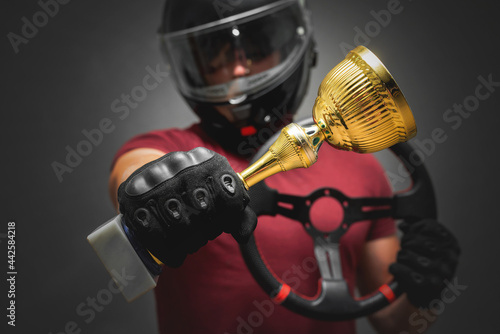 Win the auto race concept. Rally car driver in the helmet with a sport steering wheel in hand is showing a golden cup award trophy on the gray background concept. © Dmitriy