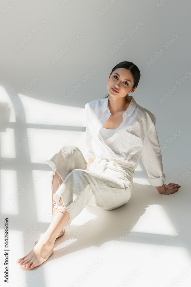 Barefoot asian woman in white clothes sitting in sunlight on grey background Barefoot asian woman in white clothes sitting in sunlight on grey background