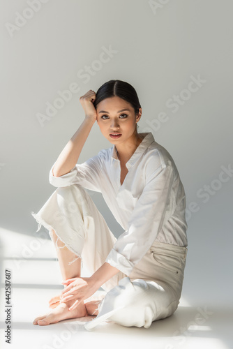 Barefoot asian woman in white clothes sitting on grey background