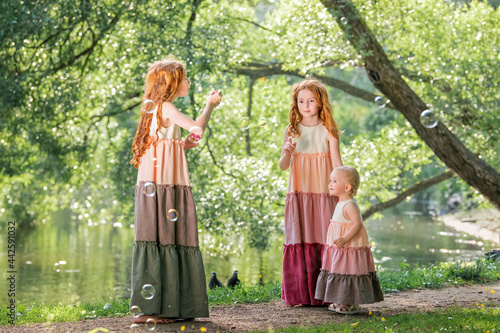 Three red-haired sisters in long linen dresses blow bubbles in the park on sunny summer day.