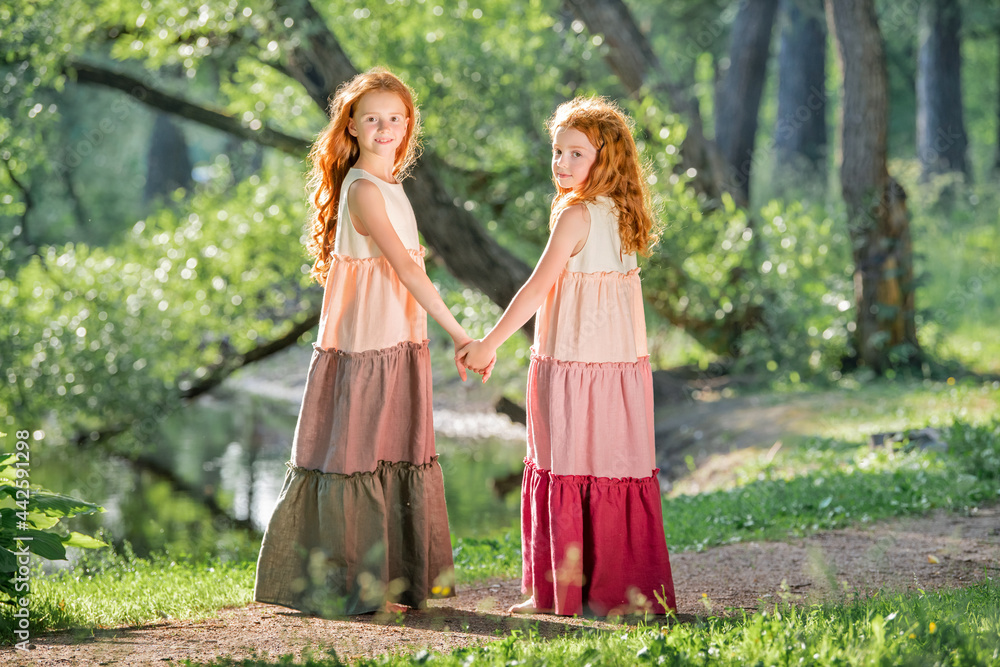 Three red-haired sisters in long linen dresses blow bubbles in the park on sunny summer day.