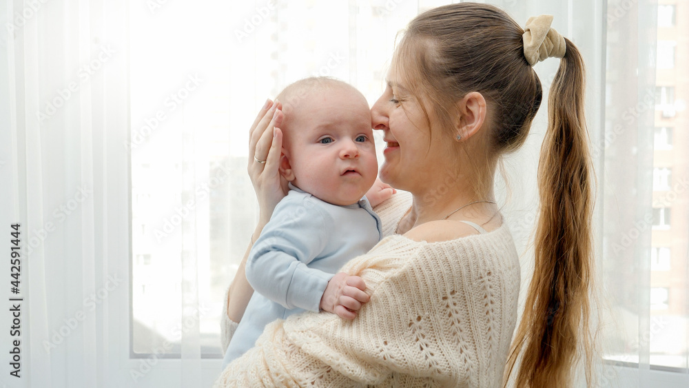 Portrait of beautiful smiling woman hugging and stroking her little baby son at big window. Concept of family happiness and child development. Portrait of beautiful smiling woman hugging and stroking her little baby son at big window. Concept of family happiness and child development.