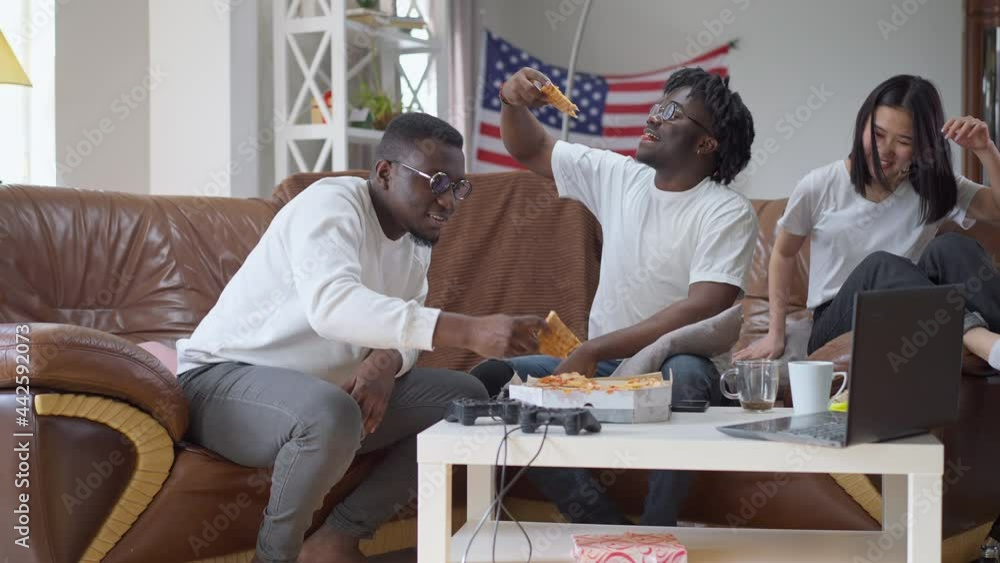 Joyful Asian woman sitting down on couch with African American men eating tasty delivered pizza. Wide shot of positive happy multiethnic friends enjoying meeting on weekend Joyful Asian woman sitting down on couch with African American men eating tasty delivered pizza. Wide shot of positive happy multiethnic friends enjoying meeting on weekend