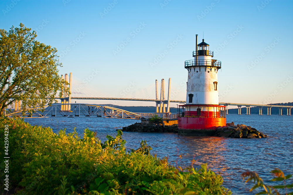 Short, red and white lighthouse on the Hudson River with a small bridge leading to it and a larger bridge further in the background -09