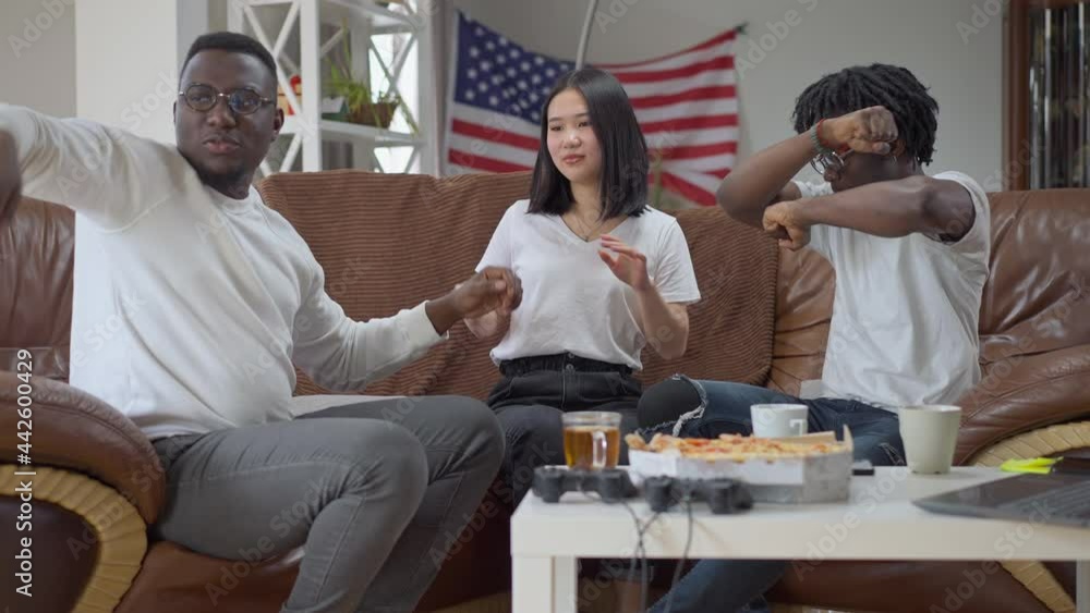 Joyful multiracial friends dancing in slow motion sitting on couch indoors. Wide shot of happy cheerful African American men and Asian woman having fun enjoying meeting on weekend Joyful multiracial friends dancing in slow motion sitting on couch indoors. Wide shot of happy cheerful African American men and Asian woman having fun enjoying meeting on weekend