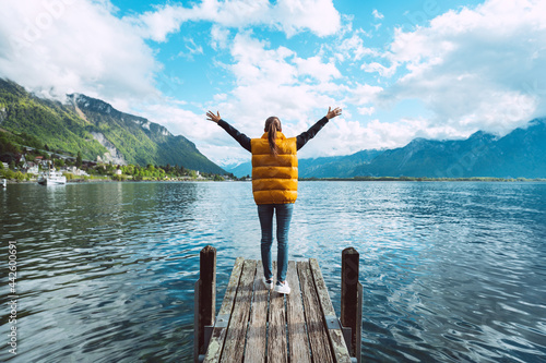 Young woman traveler with open arms standing on wooden bridge and enjoying great view on Geneva lake in Switzerland
