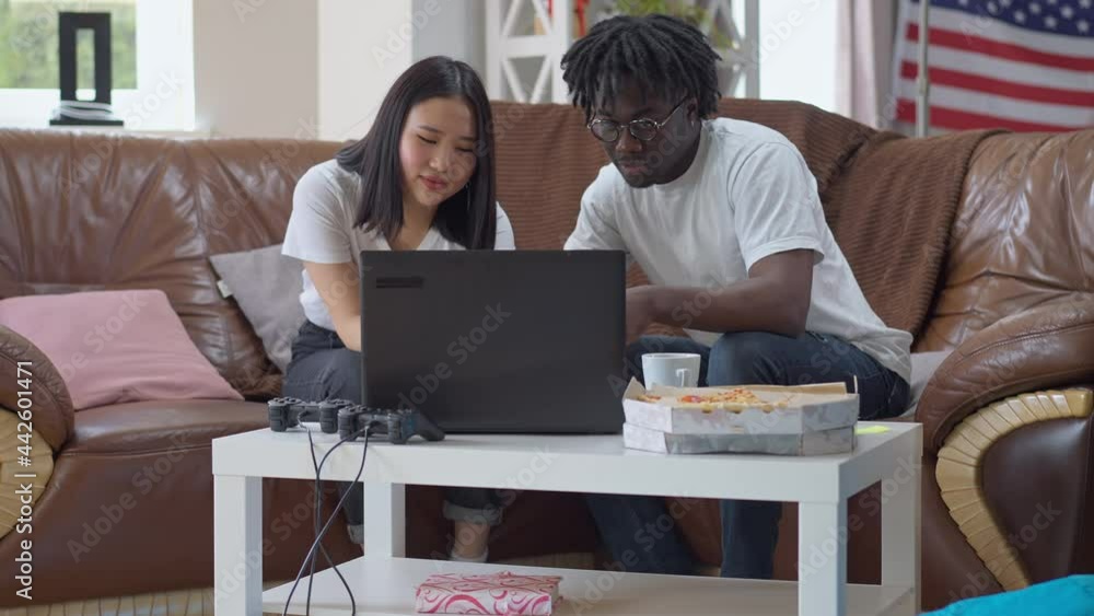 Wide shot portrait of African American man and Asian woman surfing Internet on laptop talking in slow motion. Interracial young couple enjoying weekend leisure indoors in living room Wide shot portrait of African American man and Asian woman surfing Internet on laptop talking in slow motion. Interracial young couple enjoying weekend leisure indoors in living room