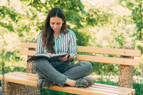 Photo of young beautiful woman sitting on wooden bench in park and writing in agenda or planner