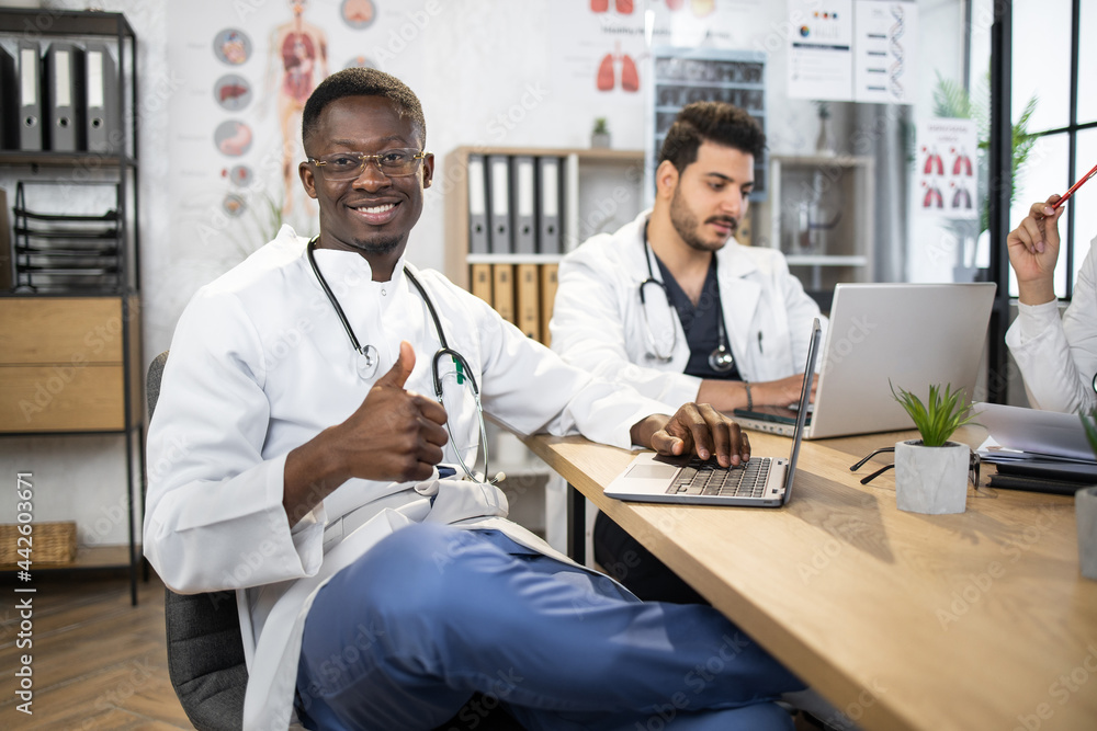 Fototapeta premium Group of competent doctors with laptops and tablets having meeting at medical office. International specialists sharing with knowledge. African male doctor shows thumb up