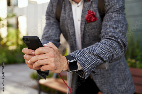 Using technologies. Close up of hands of elegant middle aged businessman using his smartphone while standing with electrical scooter outdoors in the daytime