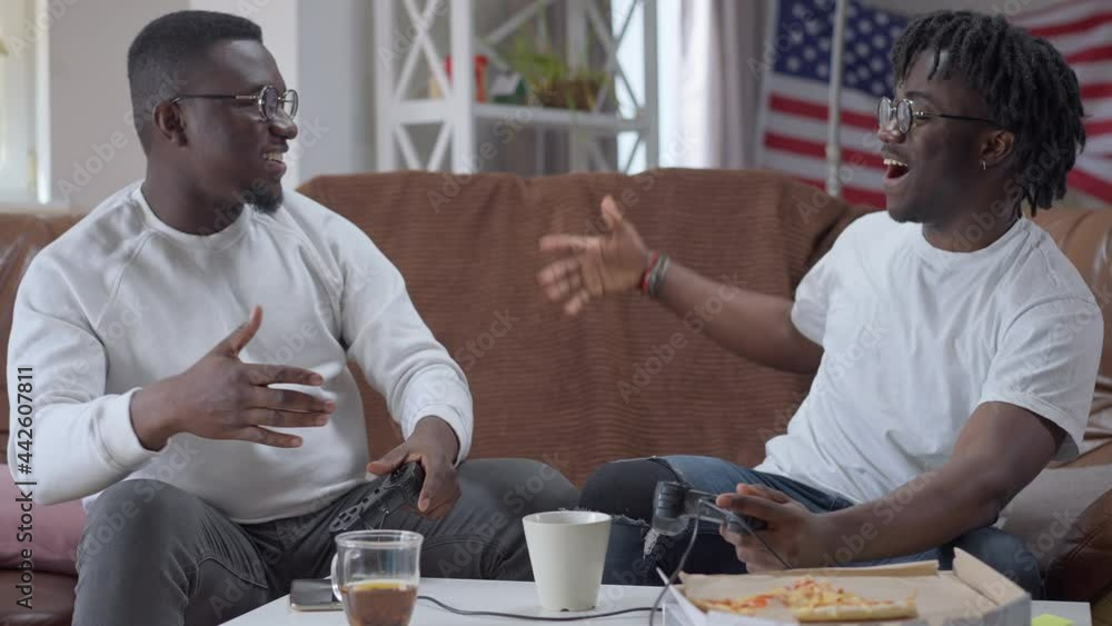 Positive millennial African American men giving high-five rejoicing victory in multiplayer video game. Happy laughing handsome friends winning resting on weekend indoors Positive millennial African American men giving high-five rejoicing victory in multiplayer video game. Happy laughing handsome friends winning resting on weekend indoors