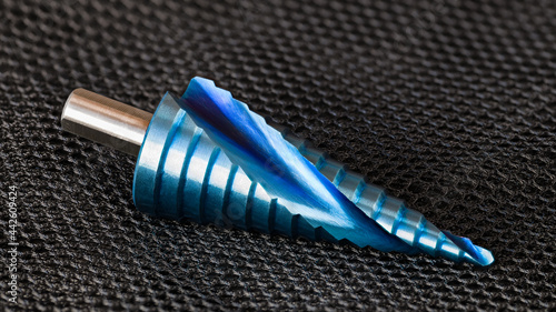 Closeup of conical step drill bit with spiral flute and blue surface finish on grid net black background. One nano coated unibit to boring varied hole sizes. Sharp metal cutting tool of quality steel. photo