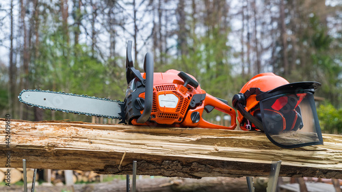 Close-up of portable chain saw and safety helmet on wood in metal sawhorse. Professional orange power chainsaw and protective hard hat with face shield of grid mesh on wooden log. Sawing machine tool. photo
