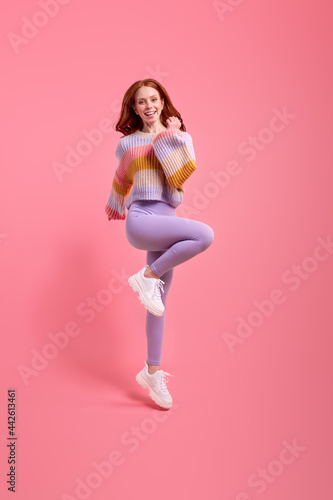 Attractive funny redhead lady jumping high raising hands first place winner wearing casual sweater, leggins isolated over bright pink color background. Beautiful lady rejoicing photo