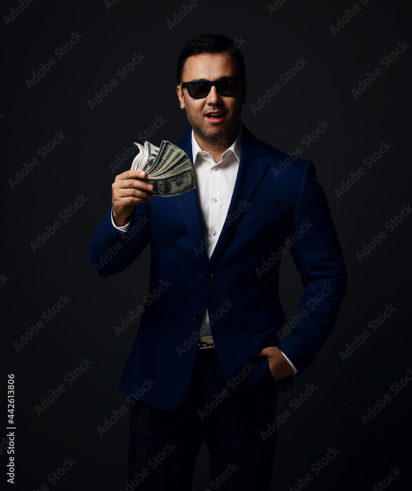 Portrait of insolent rich businessman in blue jacket, white shirt and sunglasses standing holding bundle, fan of dollars cash and the other hand in pocket of his pants over dark background Portrait of insolent rich businessman in blue jacket, white shirt and sunglasses standing holding bundle, fan of dollars cash and the other hand in pocket of his pants over dark background
