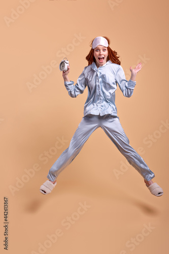 Young redhead woman wearing pajama and eye mask holding alarm clock excited, in shock, emotional expression screaming with raised hands, jumping isolated on beige background © alfa27