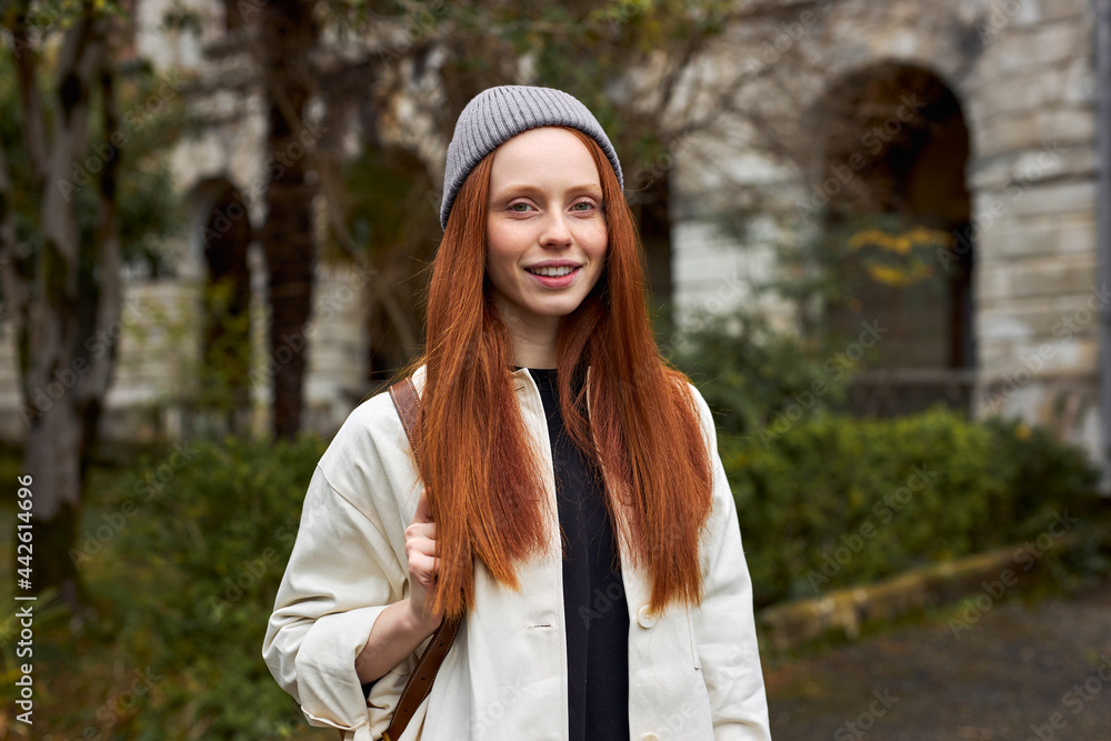 Close-up Portrait Of Young Woman With Natural Red Hair On Travel Trip, Outdoors, Posing At Camera, Smiling, In Coat And Hat. Old Building In The Background Close-up Portrait Of Young Woman With Natural Red Hair On Travel Trip, Outdoors, Posing At Camera, Smiling, In Coat And Hat. Old Building In The Background