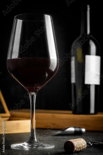 full wine glass on wooden table, with wine bottle behind, dark background and accessories on table.