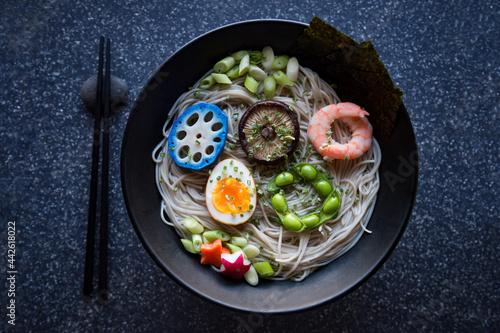 Bowl of Japanese buckwheat ramen noodle soup topped with colorful circles of blue lotus root, shiitake mushroom, shrimp, boiled egg, and edamame beans