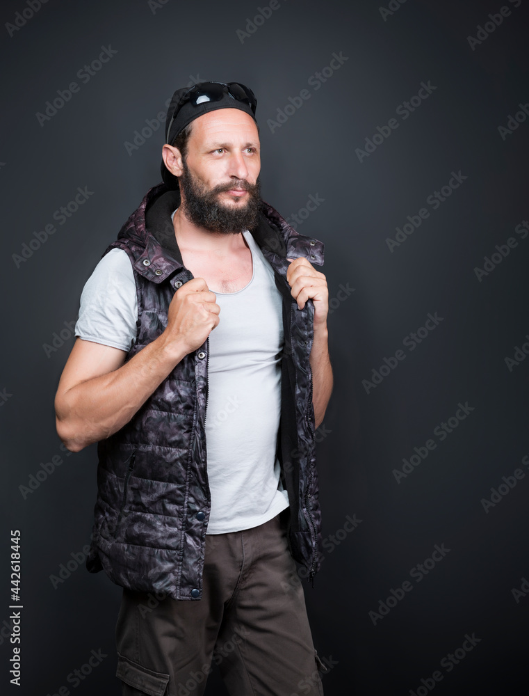 Studio portrait of american handsome bearded man wearing a cap, sunglasses, hoody vest and jeans, urban style or sport casual. Serious Russian Man with open vest on with hood looking away to side